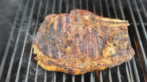 Grilled beef steak with char marks on a smoky black grill.