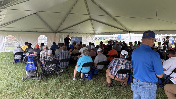 Group of people seated under a white tent on grassy ground, listening to a speaker.