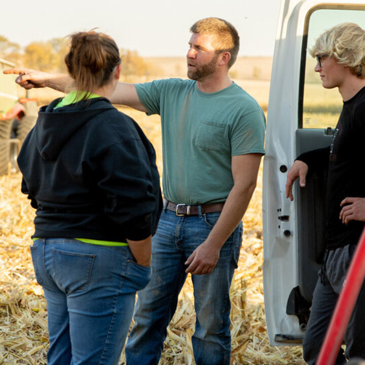 Three people stand talking in a cornfield near a parked truck and green farming equipment.